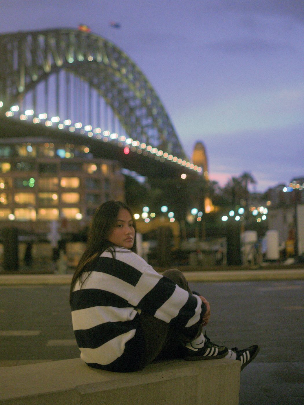 Karmina Mae seated by Sydney Harbour at dusk, the Harbour Bridge lit behind her